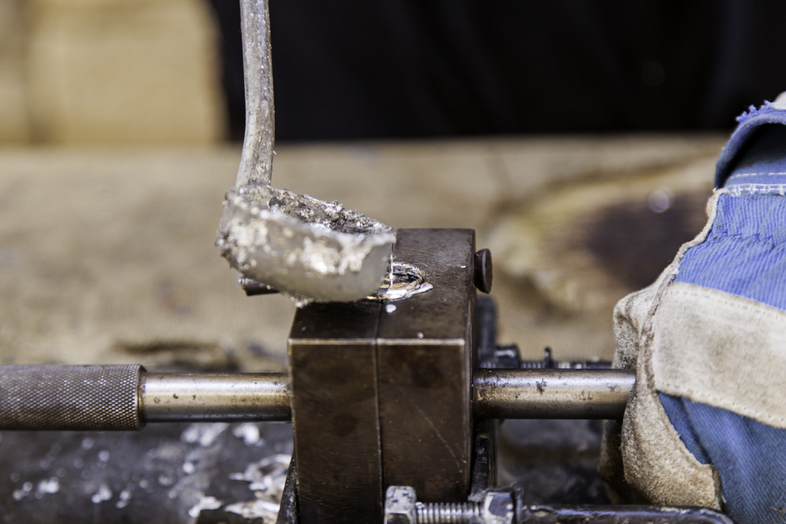 Close-up of a metalworking process, showing a worker using a tool to shape molten metal in a workshop. The image highlights the precision and craftsmanship involved in metal fabrication, with tools and equipment visible in the background.