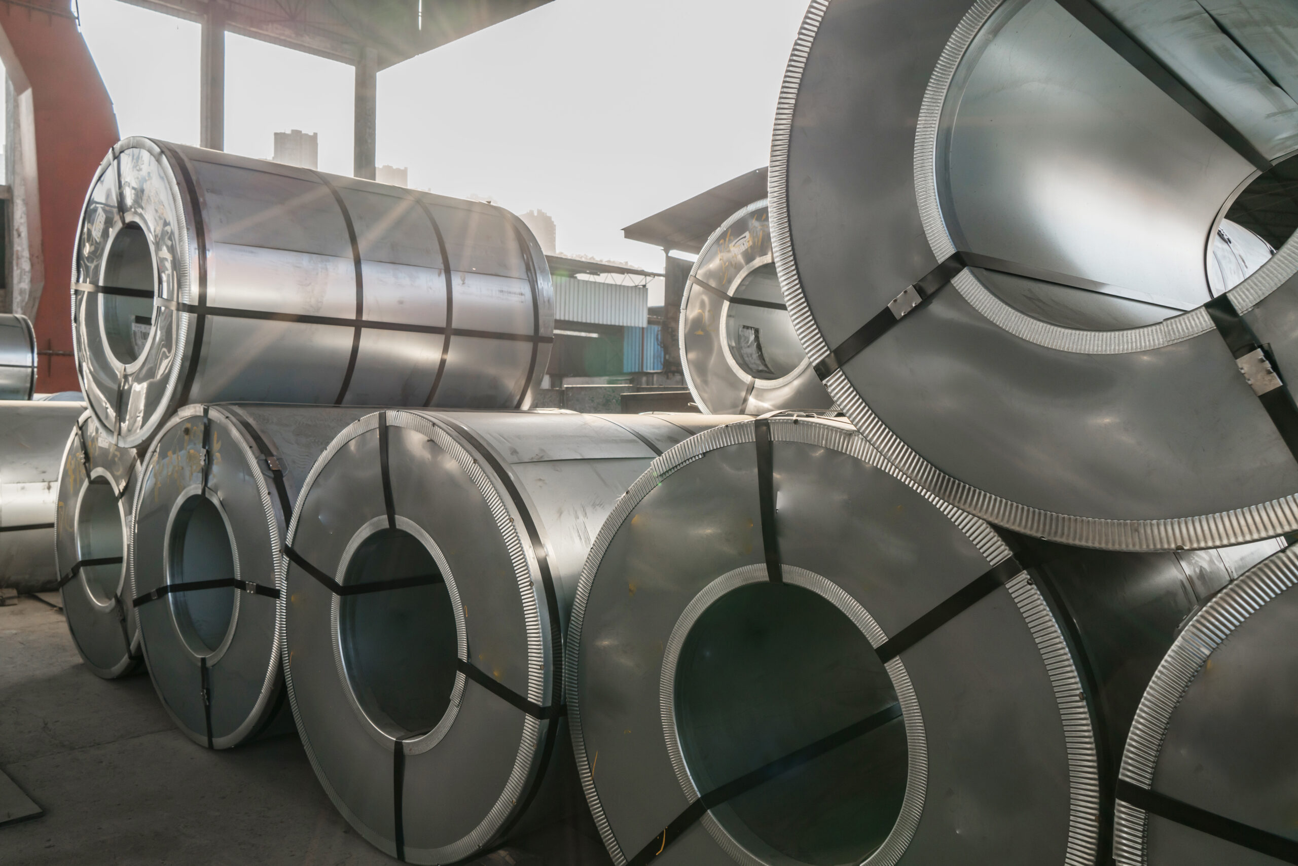 Stacks of large metal coils stored in a warehouse, showcasing their shiny surfaces and circular openings, with natural light illuminating the industrial setting.
