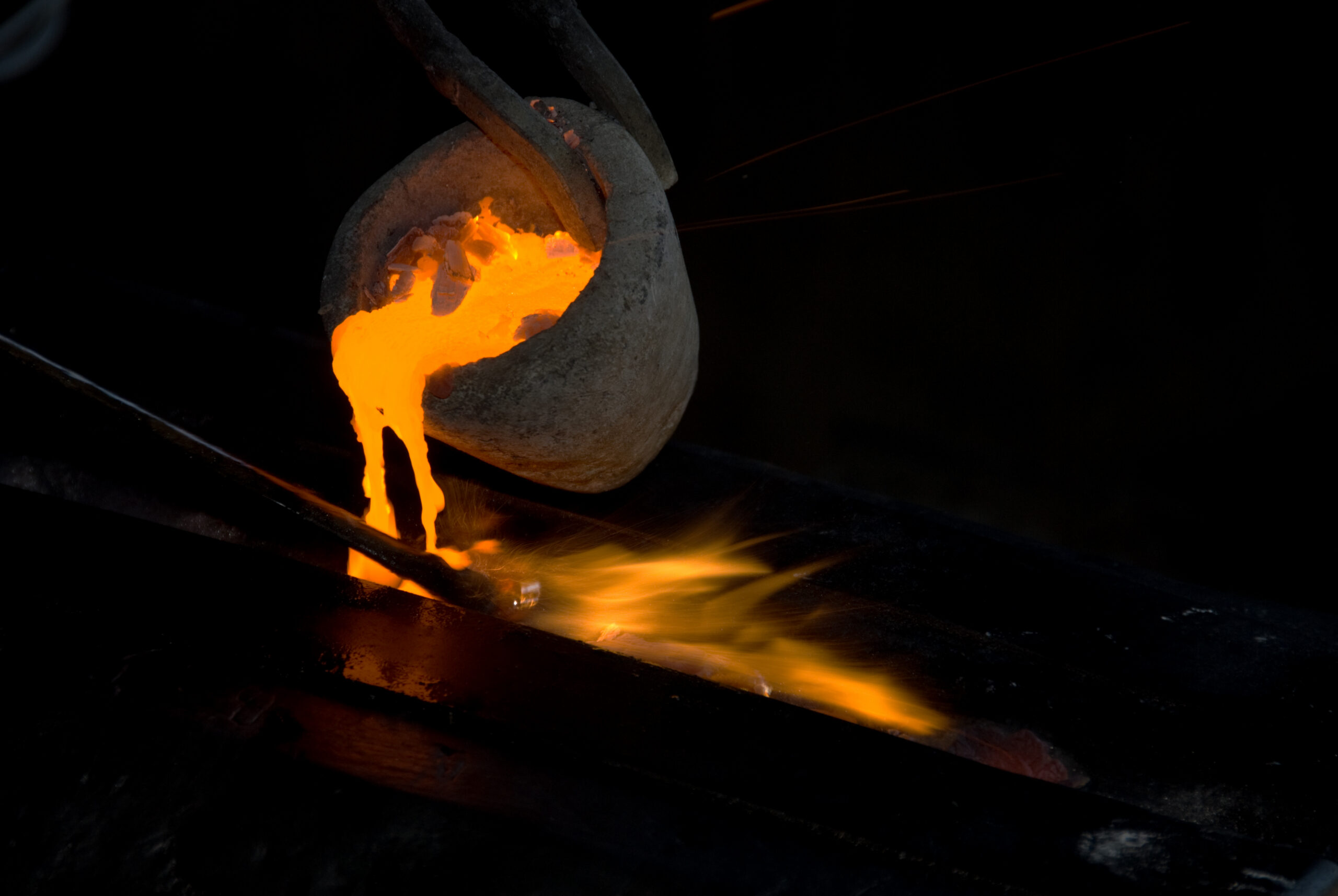Molten metal being poured from a crucible, glowing orange and creating sparks as it flows onto a black surface, showcasing the process of metal casting in a foundry.