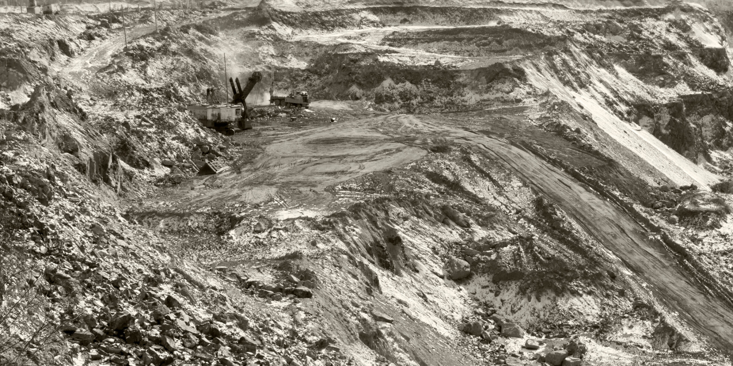 Aerial view of an open-pit mining site featuring heavy machinery, rocky terrain, and steep slopes in a monochrome setting, illustrating the impact of mining operations on the landscape.