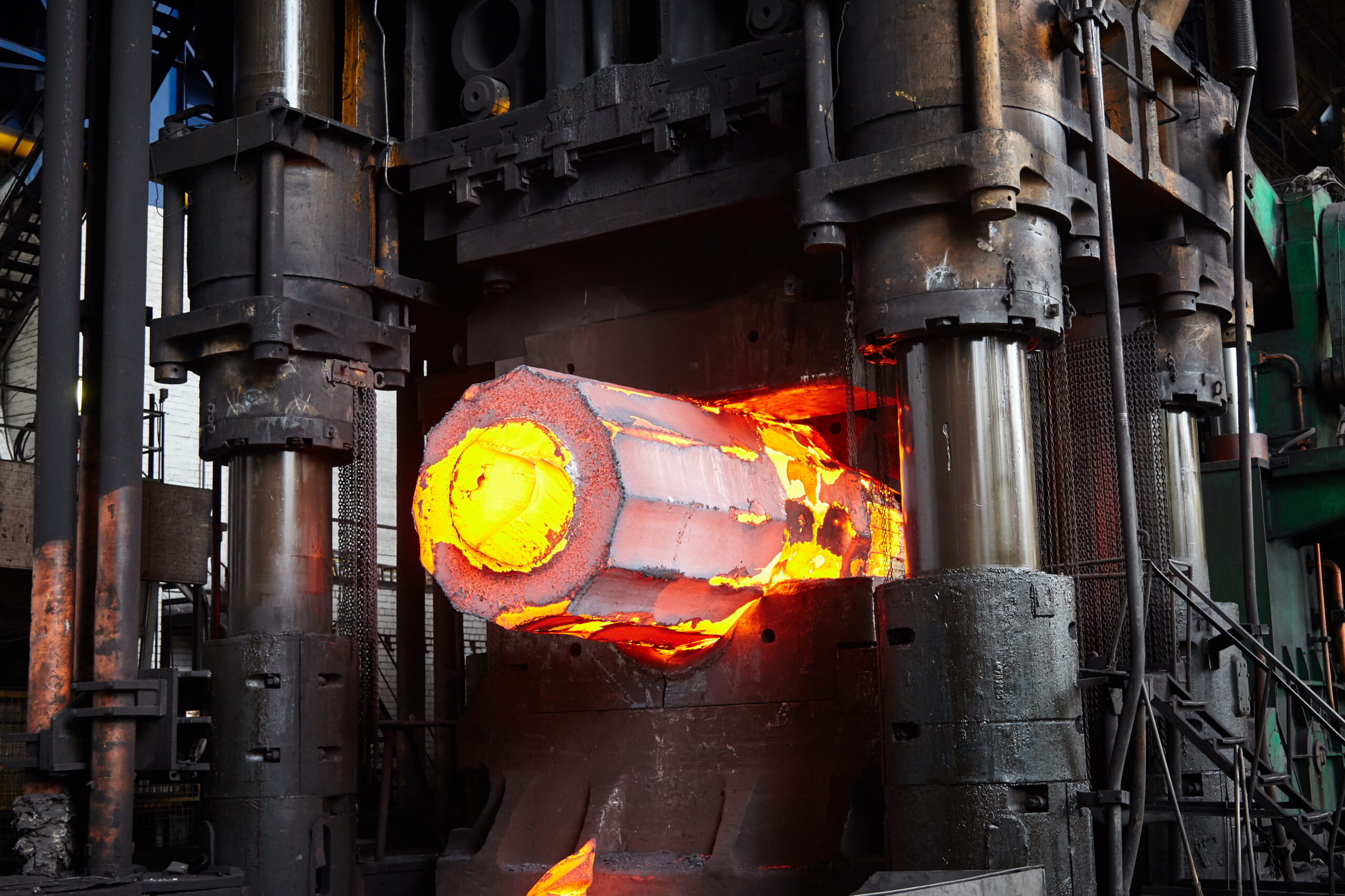 Industrial forging process showing a heated metal billet being shaped by a hydraulic press in a manufacturing facility. The vibrant orange glow of the molten metal highlights the intense heat and energy involved in metalworking.