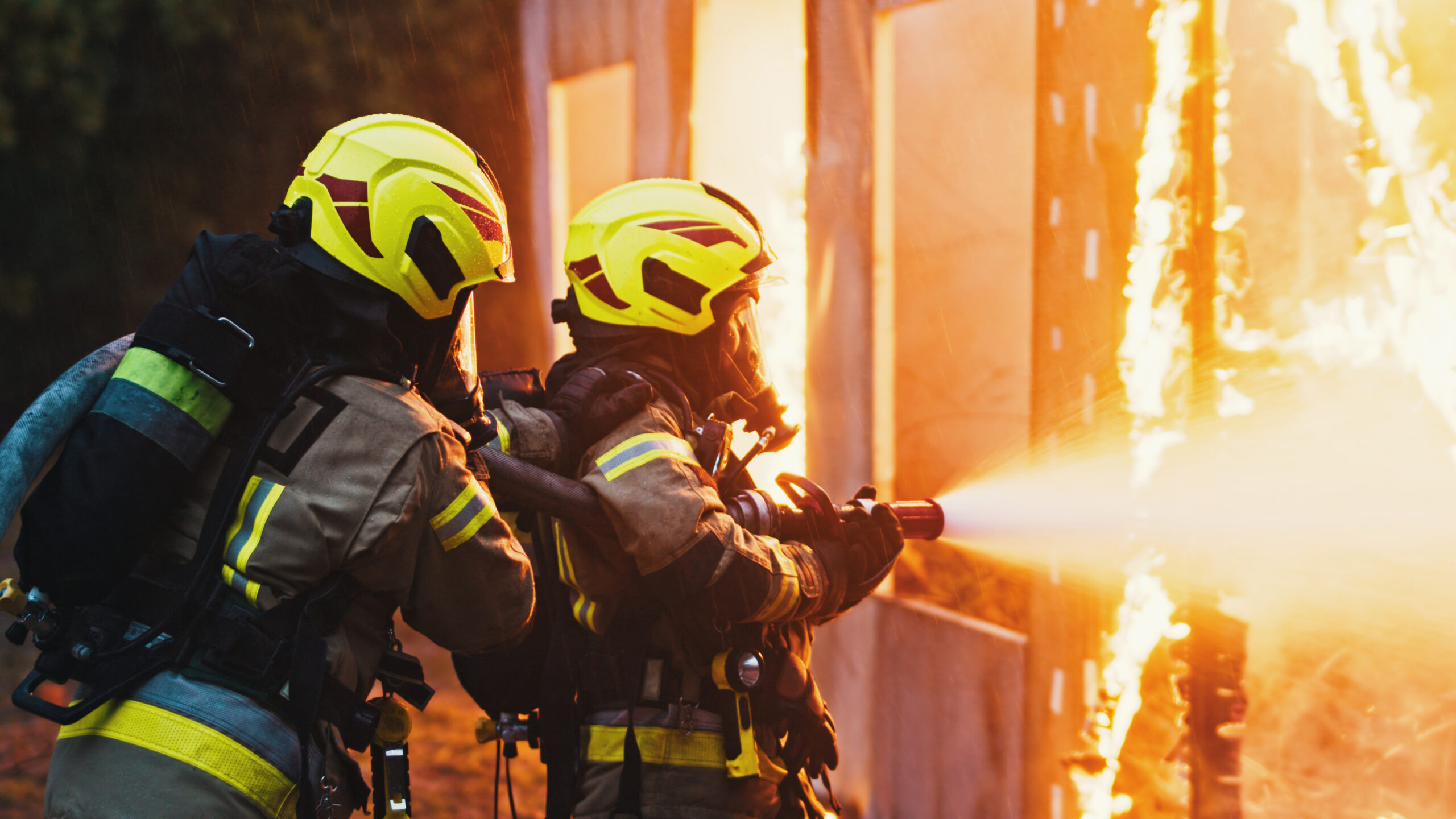 Firefighters in protective gear extinguishing flames with a hose, showcasing teamwork and emergency response during a fire incident.