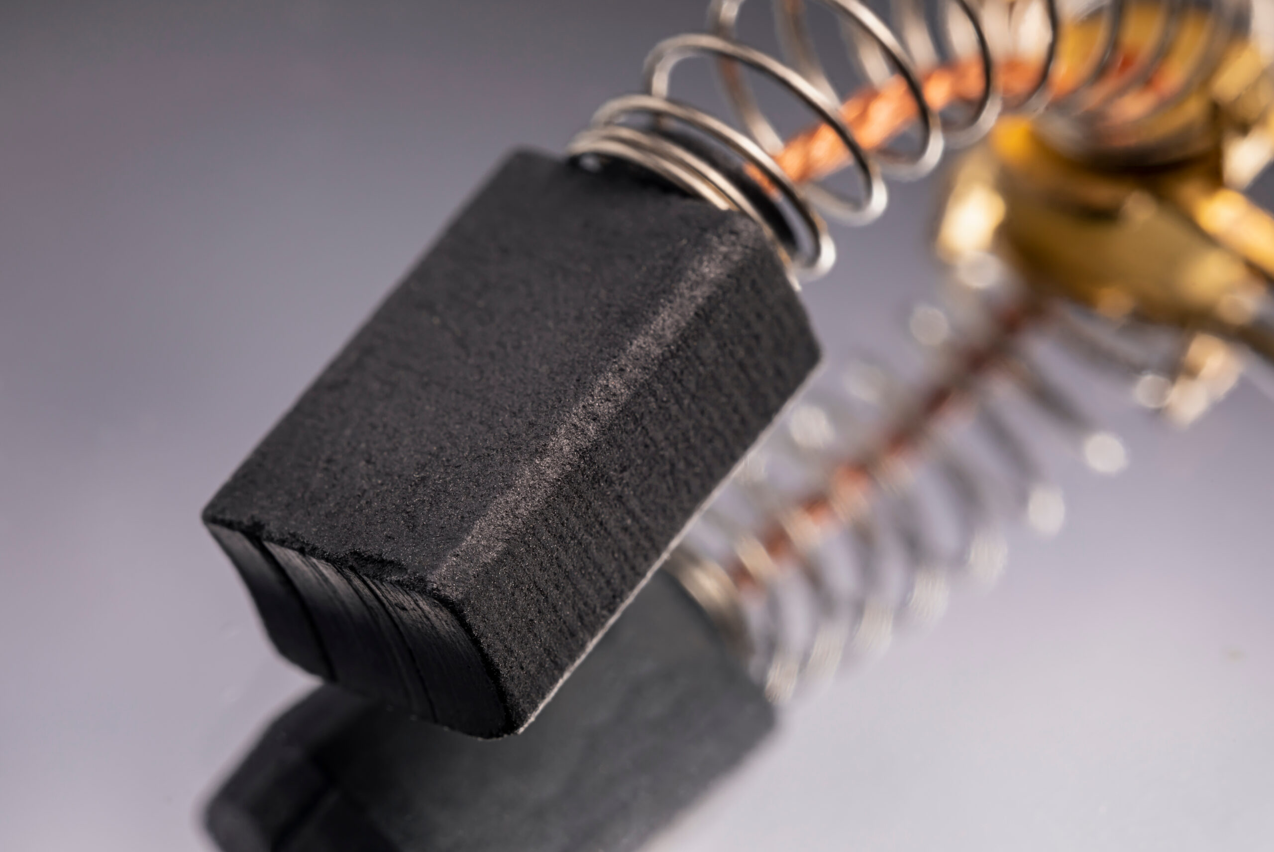 Close-up image of a carbon brush with a metallic spring, often used in electric motors and generators for conducting electricity. The brush is shown resting on a reflective surface, highlighting its texture and shape.