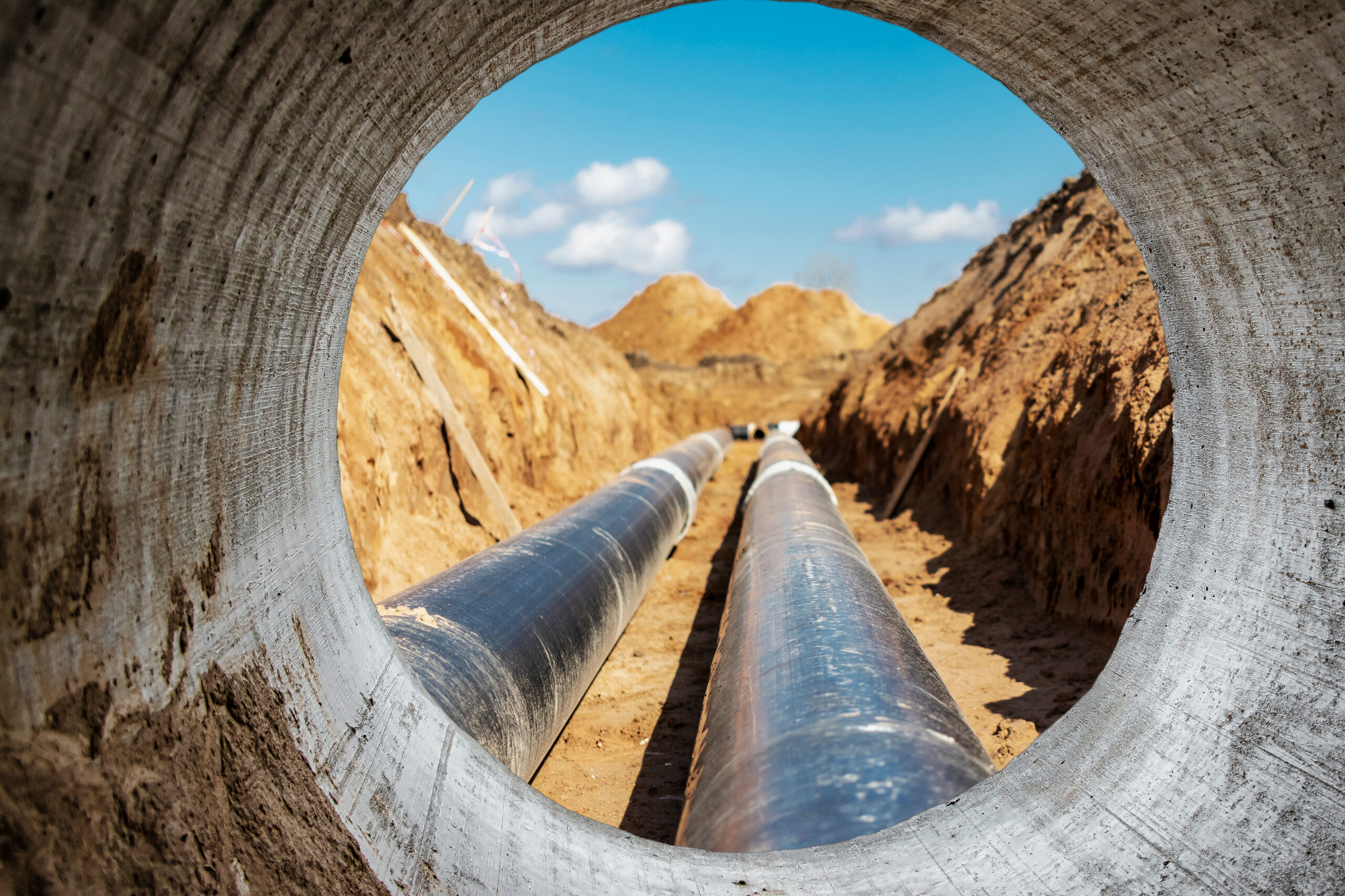 View of underground pipes through a concrete tunnel, showcasing a construction site with exposed earth and machinery in the background. The image highlights the installation of black pipelines in a trench, under a clear blue sky.