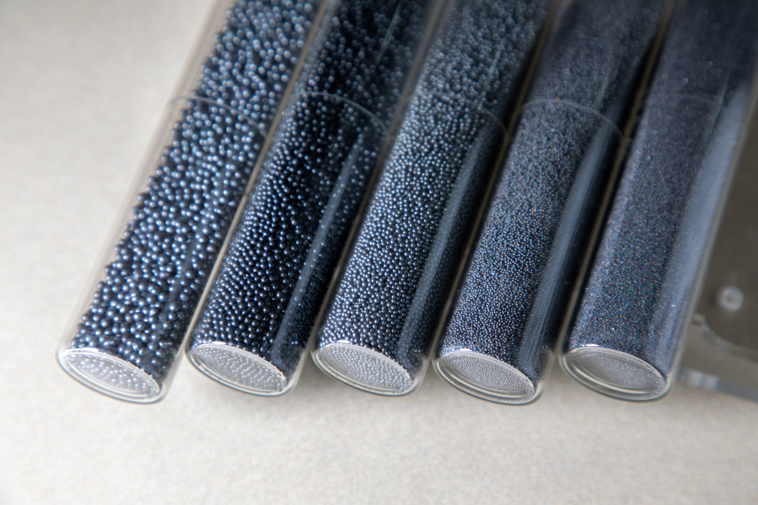 Close-up image of five transparent test tubes filled with varying sizes of black spherical beads, arranged in a row on a light surface, showcasing different textures and finishes of the beads.