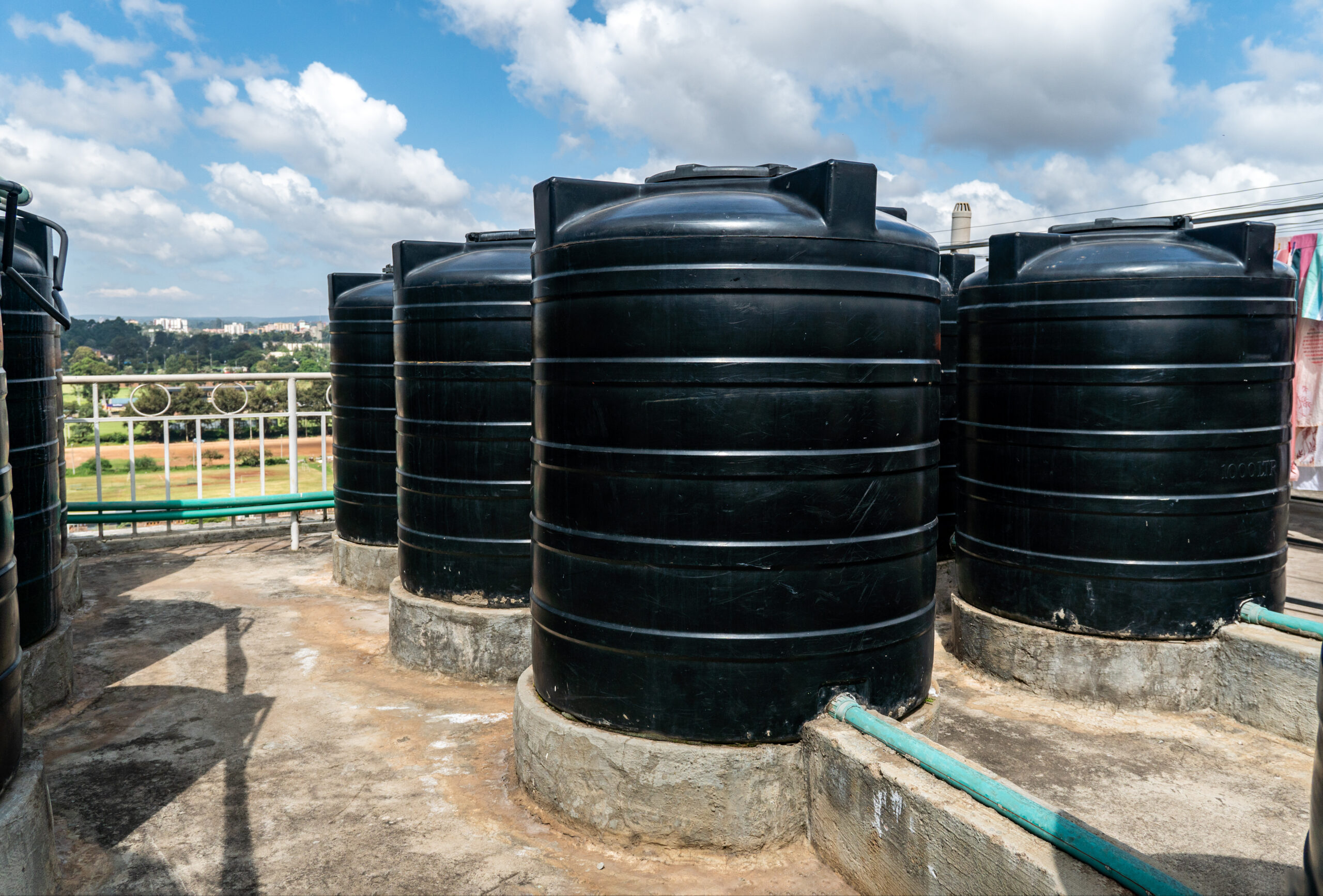 Black water storage tanks on a rooftop with a scenic view of the landscape and blue sky. The tanks are positioned on a concrete base, showcasing a modern water conservation system.