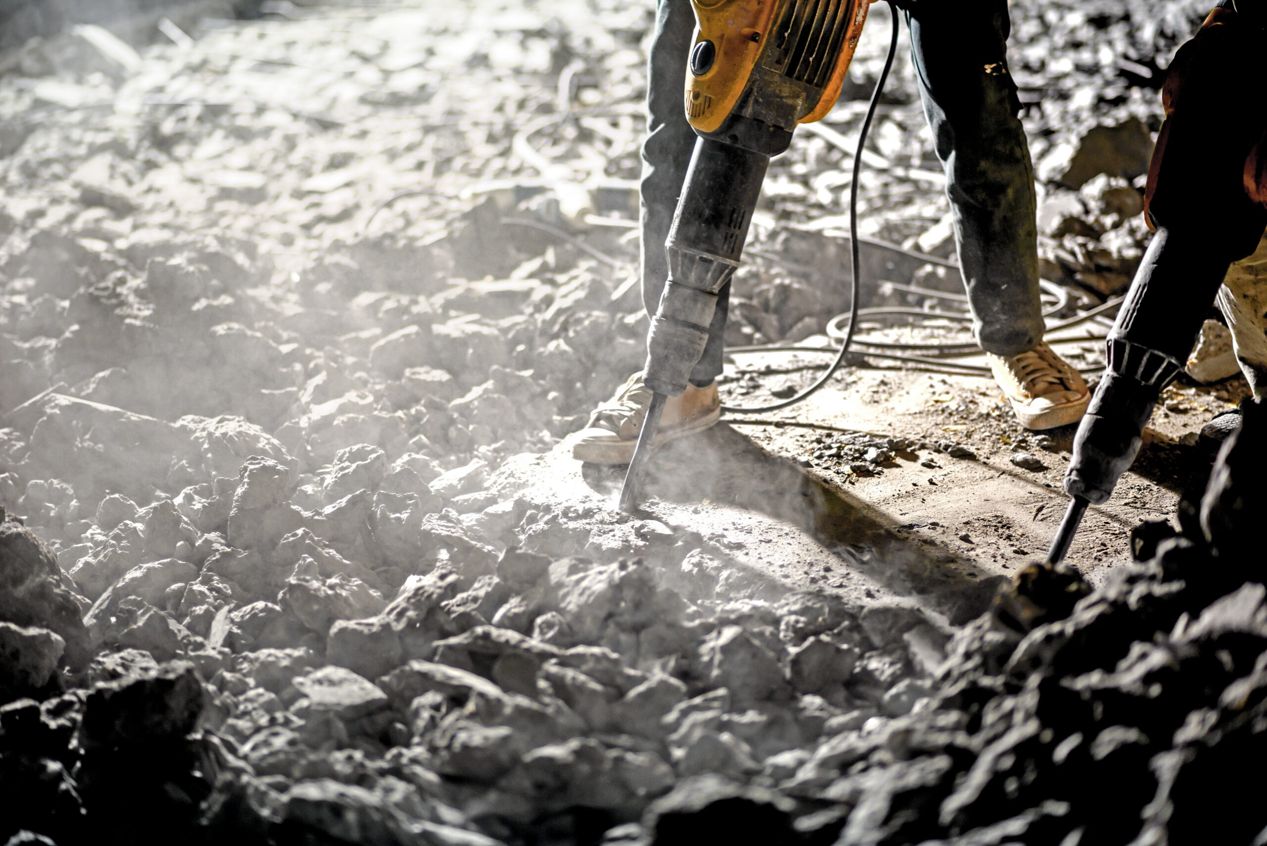 Construction workers using pneumatic jackhammers to break concrete on a rubble-strewn site, with dust and debris in the air.