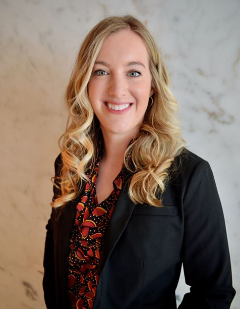 Professional headshot of a smiling woman with long, wavy blonde hair, wearing a black blazer over a colorful patterned blouse, set against a light marble background.