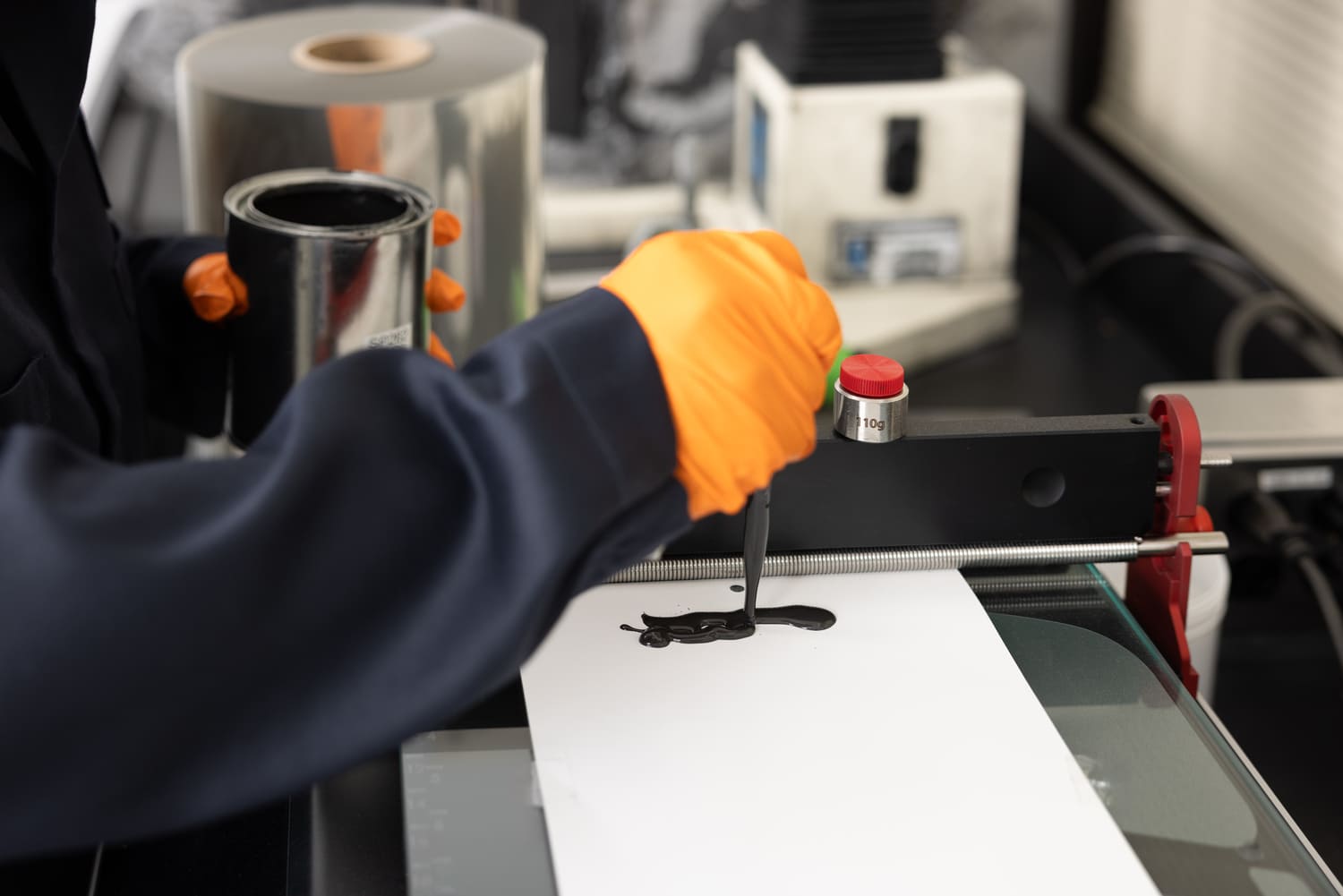 Person in a lab coat and orange gloves using a spatula to spread black ink on a sheet of paper, with printing equipment and materials visible in the background.