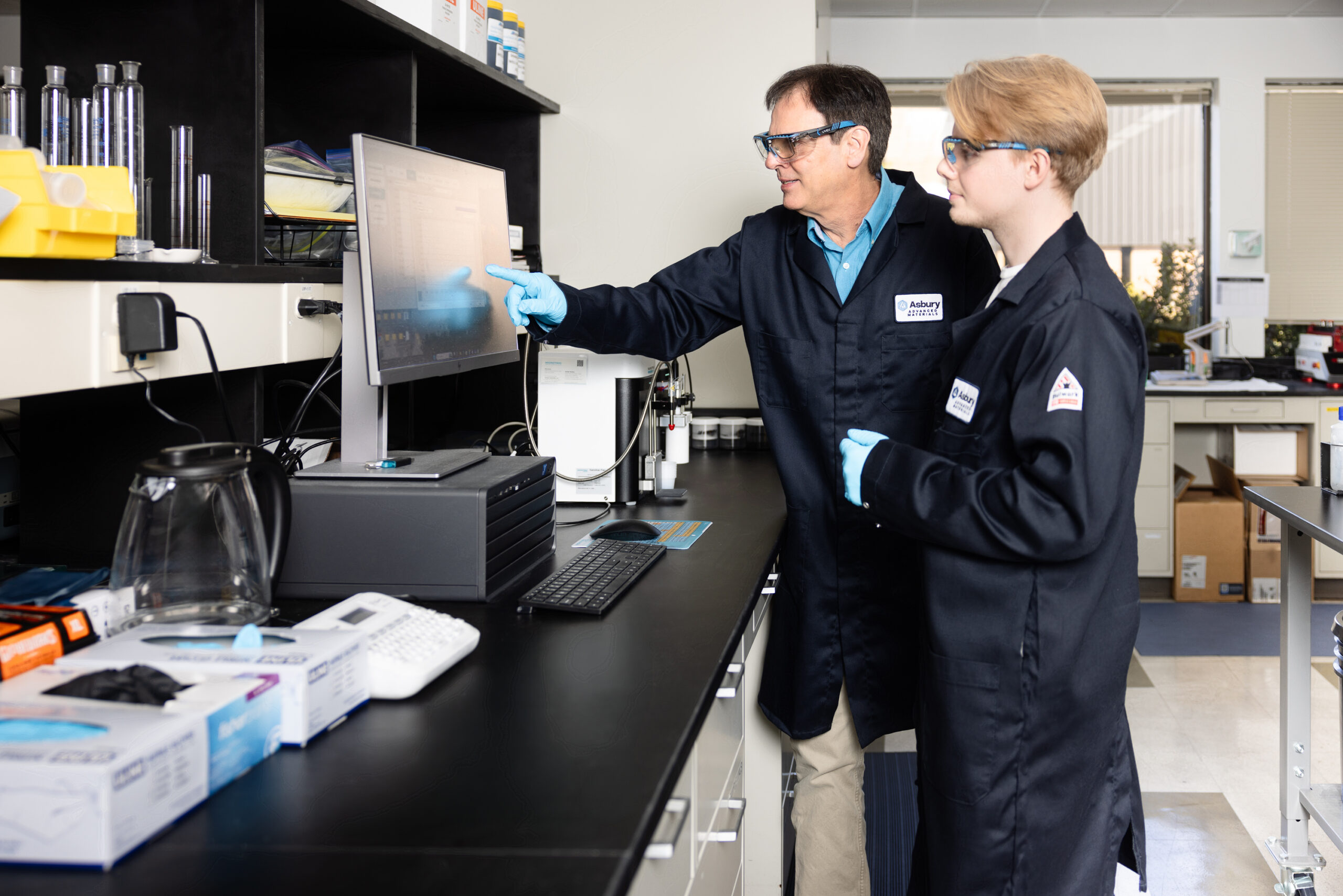 Two professionals in lab coats and gloves collaborate at a computer in a laboratory setting. One person points at the screen while the other observes, surrounded by lab equipment and supplies. The environment highlights a focus on scientific research and education.