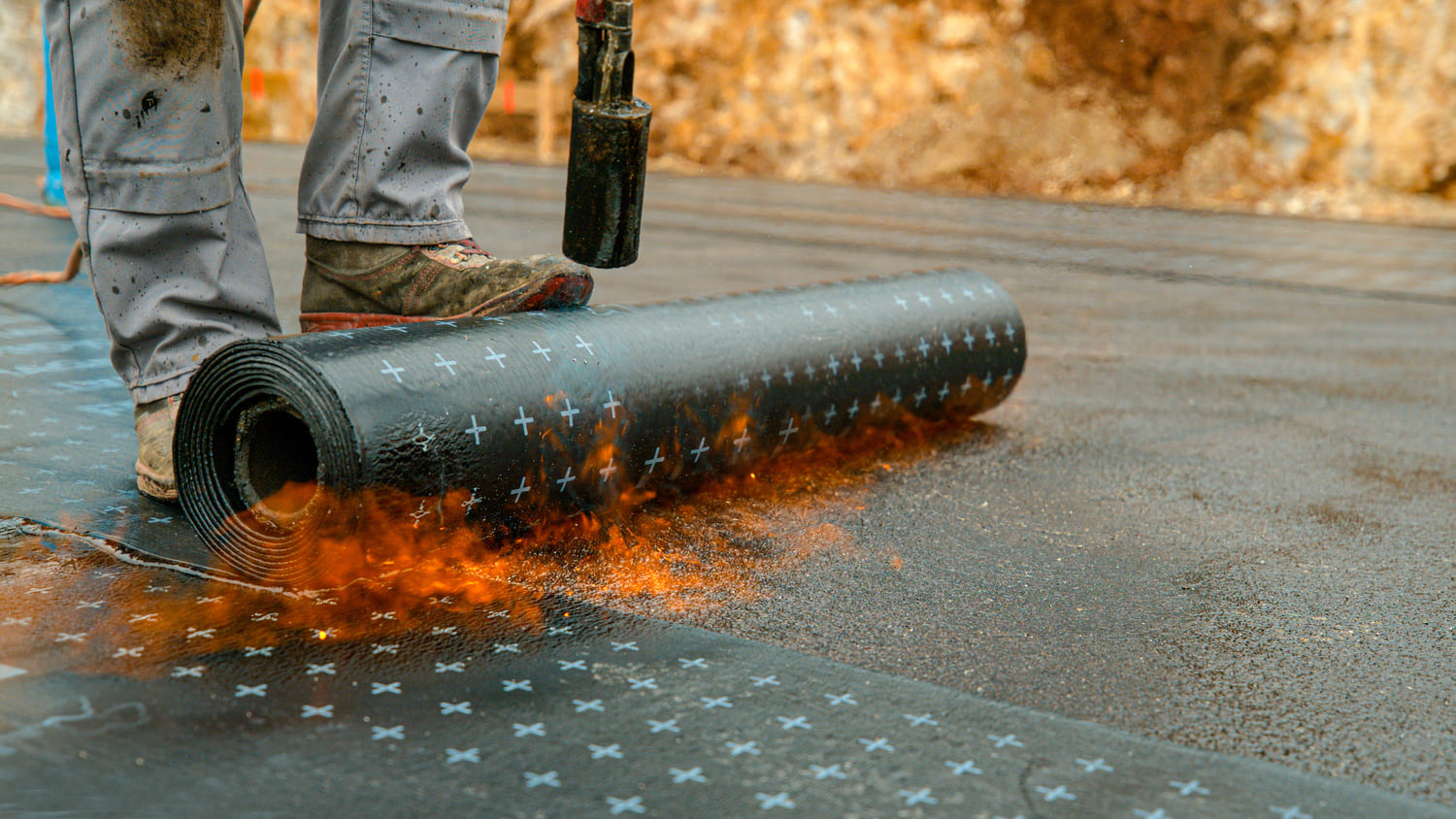 Worker applying a heat-welded roofing membrane with a torch on a construction site, showcasing the process of installing waterproof roofing materials.