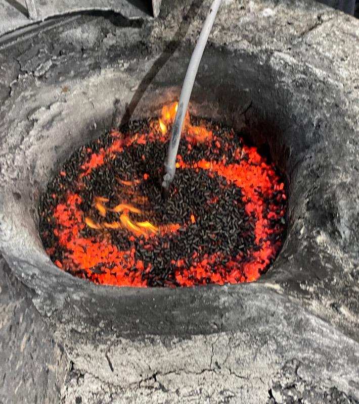 Close-up of a traditional charcoal furnace with glowing red embers and blackened charcoal, showcasing flames and a heating element in the center, used for metalworking or cooking.