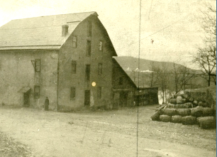 Historic rural building with a large barn and haystack in the foreground, surrounded by trees and a dirt road, showcasing early 20th-century architecture.