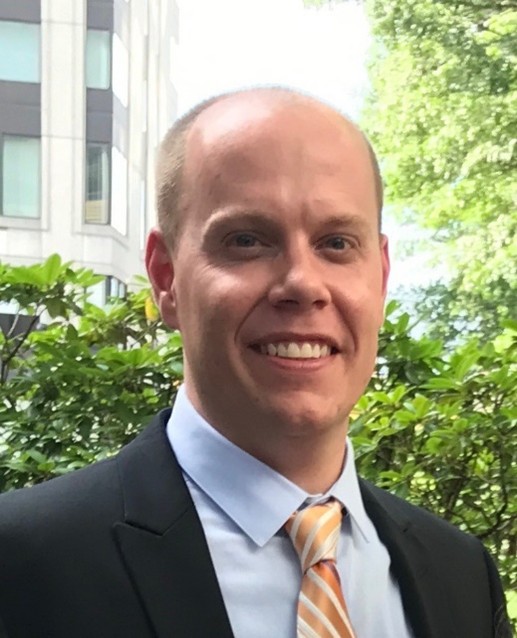 Smiling man in a business suit with a light blue shirt and orange-striped tie, standing outdoors with greenery and a modern building in the background.