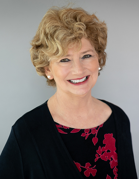 Smiling woman with curly blonde hair wearing a black and red floral blouse, posing against a neutral gray background. Ideal for professional profiles or business-related content.