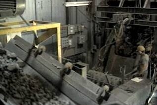 Industrial setting featuring machinery and a conveyor system transporting materials, with a worker in safety gear operating equipment in the background.