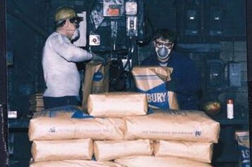 Two workers in protective gear handling large bags of a material in a warehouse setting. One worker is operating machinery while the other prepares bags for transport. The environment appears industrial, with visible electrical equipment in the background.