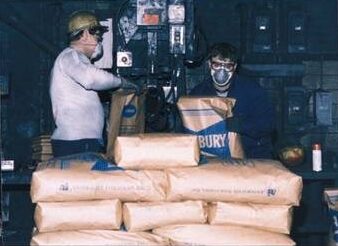 Workers handling bags of natural flake graphite in a manufacturing facility, showcasing safety measures with masks and helmets.
