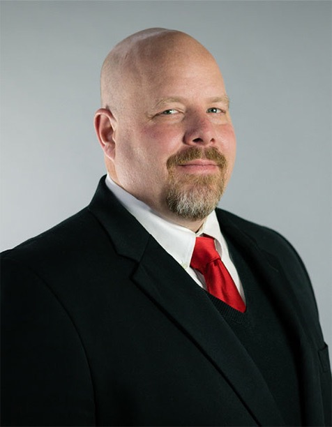 Professional headshot of a bald man wearing a black suit and a red tie, smiling confidently against a neutral gray background.