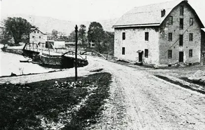 Black and white historical image of the Graphite Mill along the Musconetcong River in Asbury, New Jersey, featuring a dirt road leading to the mill and surrounding buildings.