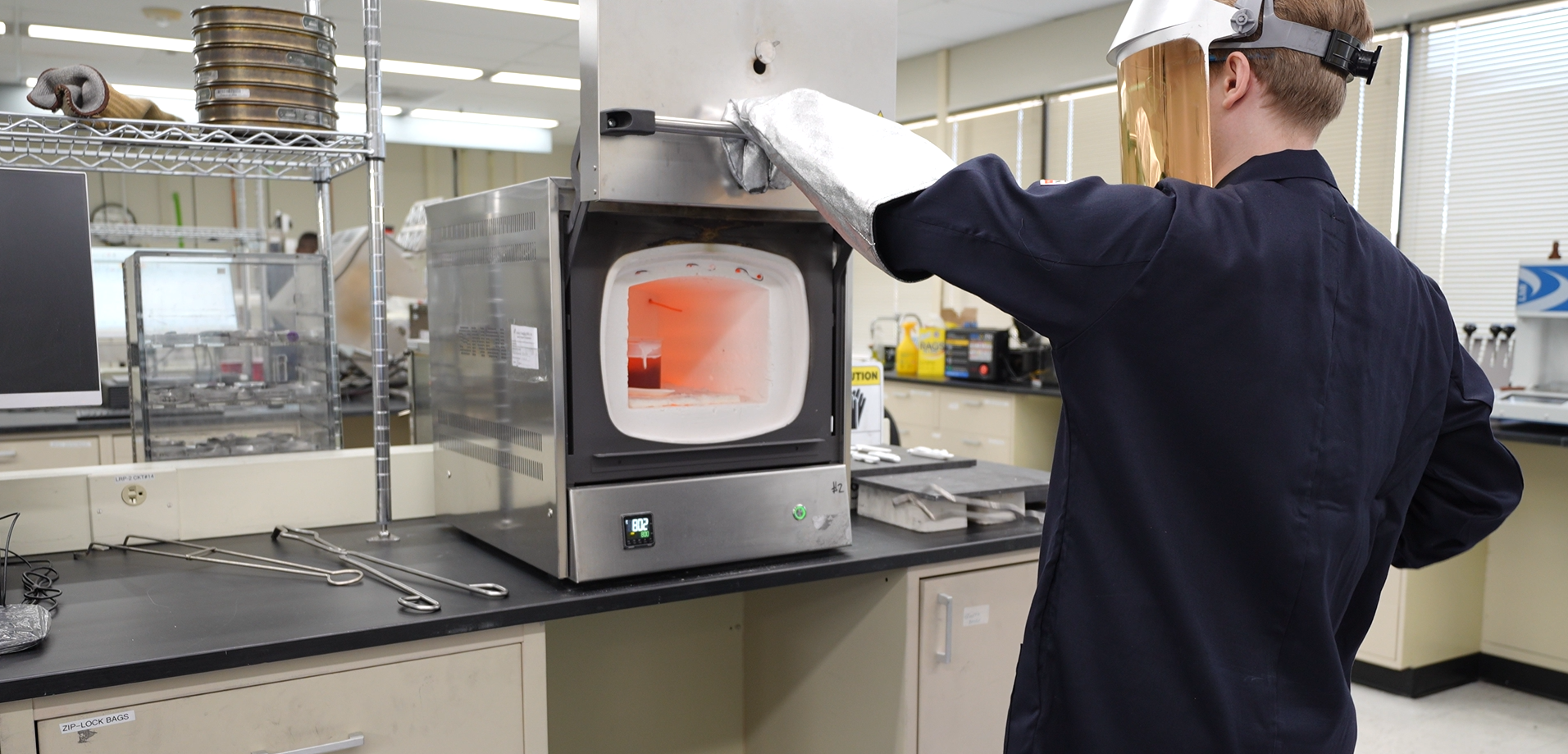 Laboratory technician operating a high-temperature furnace, wearing protective gear, in a research lab setting. The image showcases safety protocols and equipment used for material testing and analysis.
