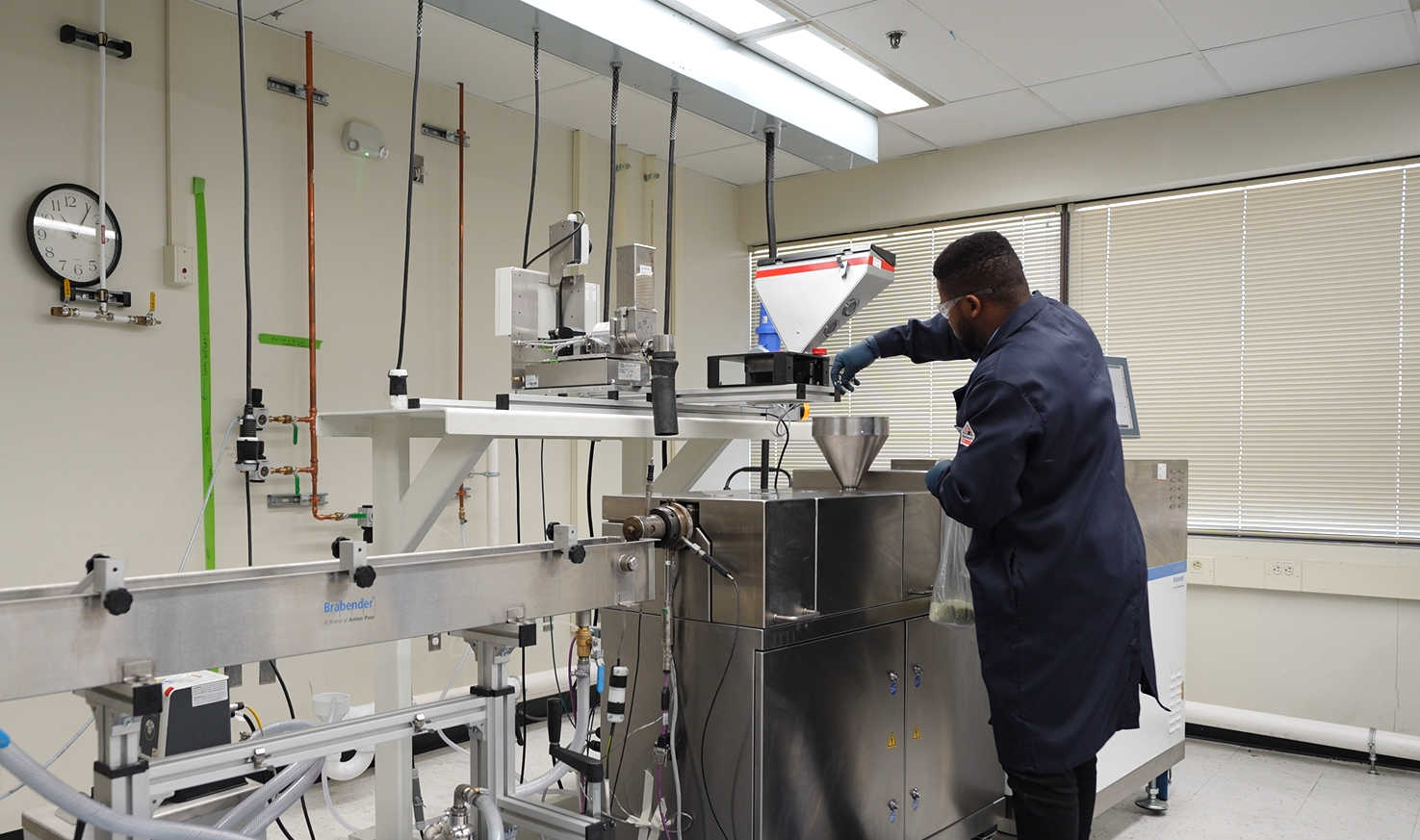 Laboratory technician operating advanced processing equipment in a research lab, focusing on materials handling and measurement with a funnel and digital display. The environment features safety equipment and organized workspace elements.