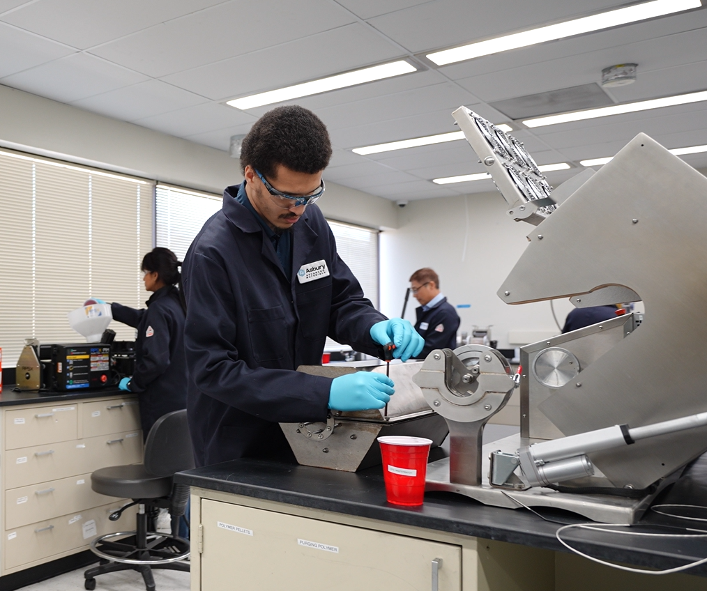 Students conducting experiments in a modern laboratory, one male student wearing safety goggles and gloves is working on a machine, while a female student in the background is pouring a liquid into a container. Red cups and laboratory equipment are visible on the counter.