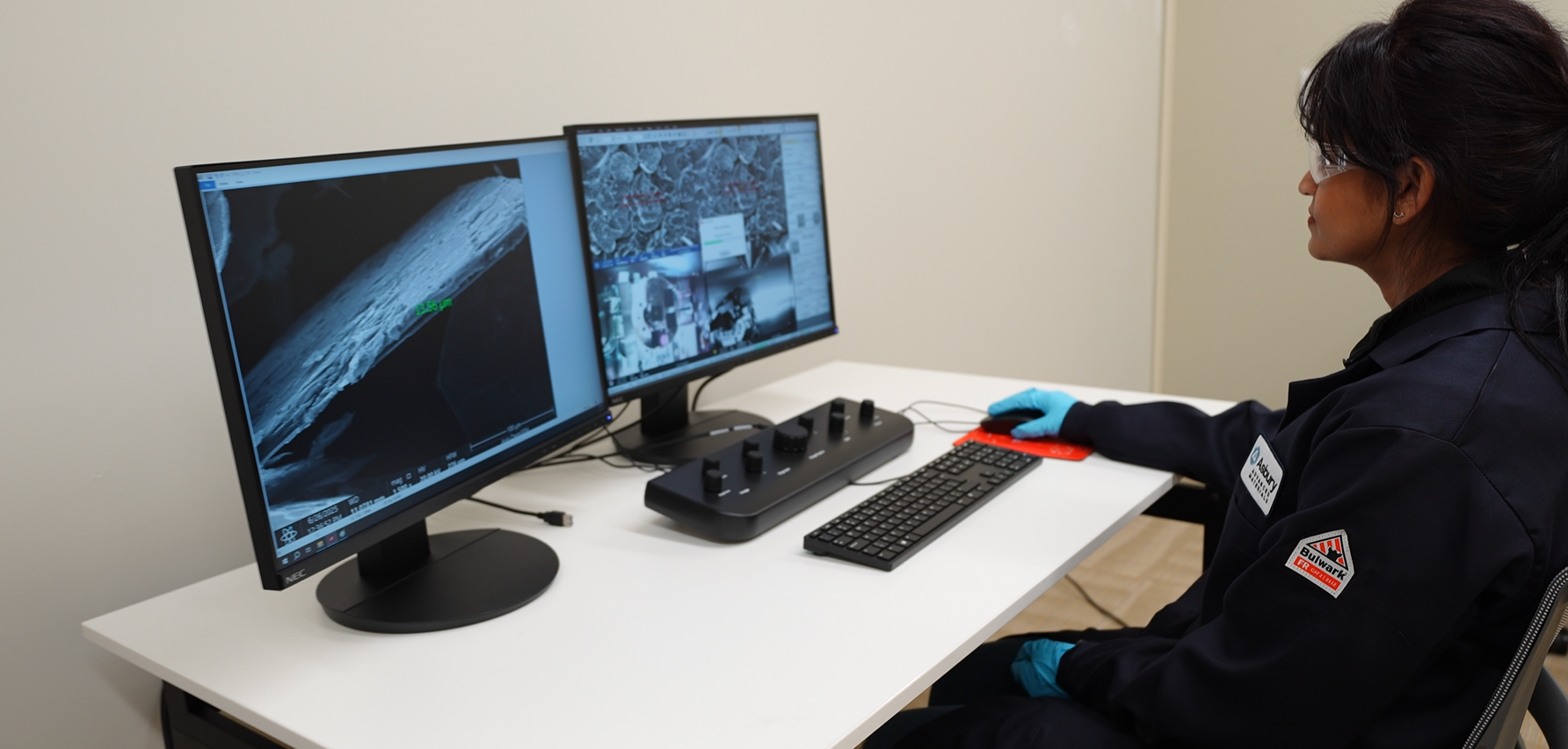 Scientist analyzing microscopic images on dual monitors in a laboratory setting, with focus on detailed surface structures and measurements, wearing protective gloves and eyewear.