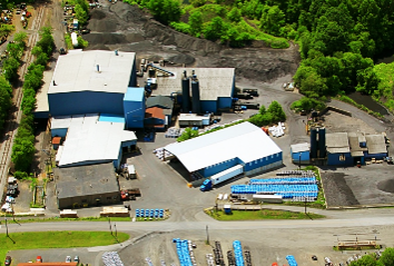 Aerial view of a manufacturing facility with multiple buildings, surrounded by greenery and industrial equipment, showcasing a mix of storage areas and processing plants.