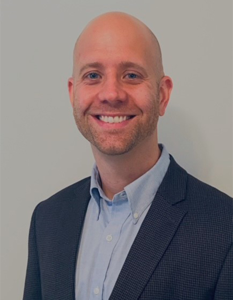 Smiling professional man in a suit and collared shirt, standing against a neutral background, representing business and corporate environments.