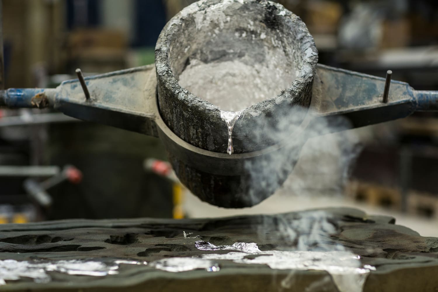 Molten metal pouring from a crucible into a mold, with steam rising in a metalworking workshop, illustrating the casting process in metal fabrication.