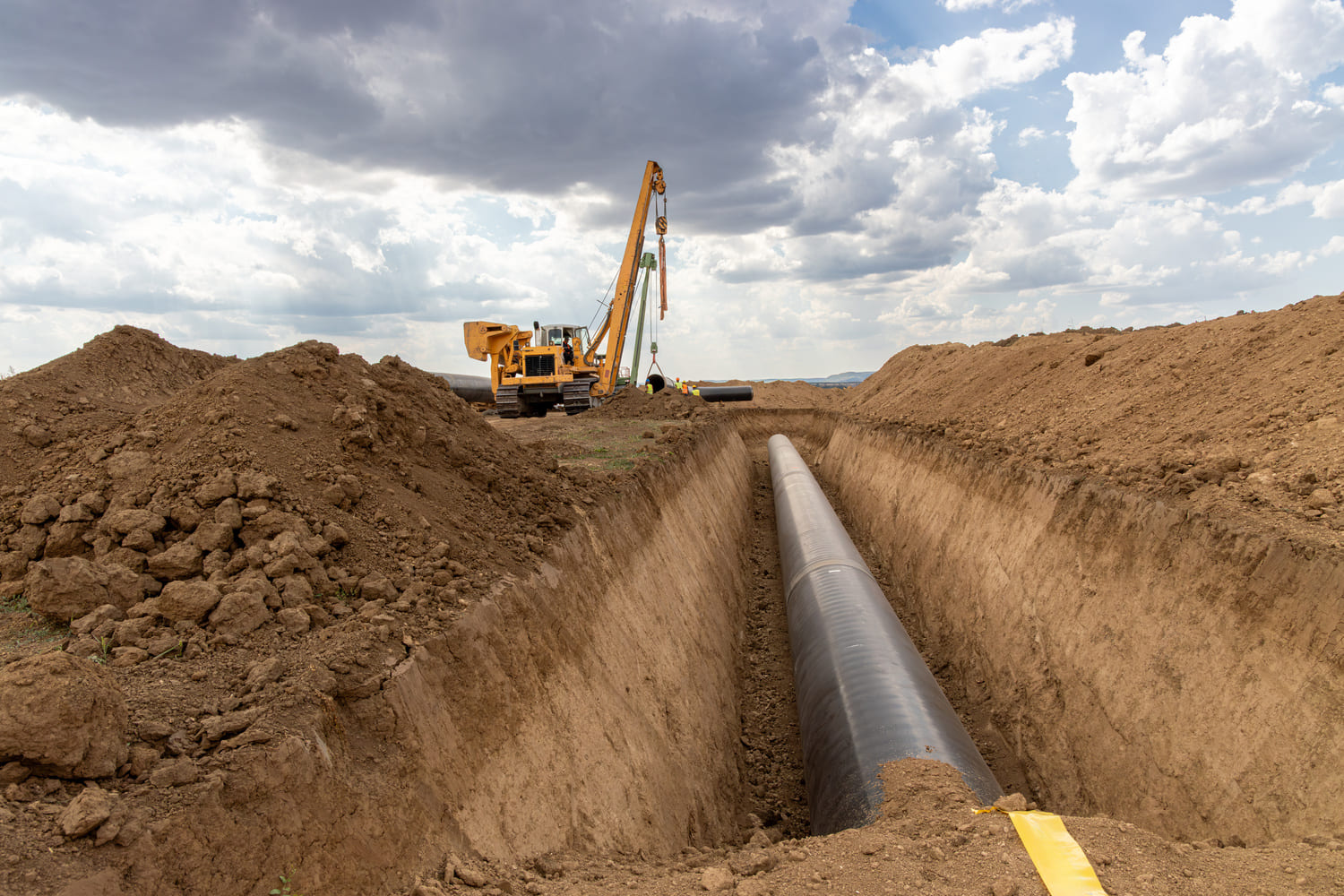 Construction site featuring a large trench with a black pipeline being installed, surrounded by piles of excavated soil and heavy machinery in the background under a cloudy sky.