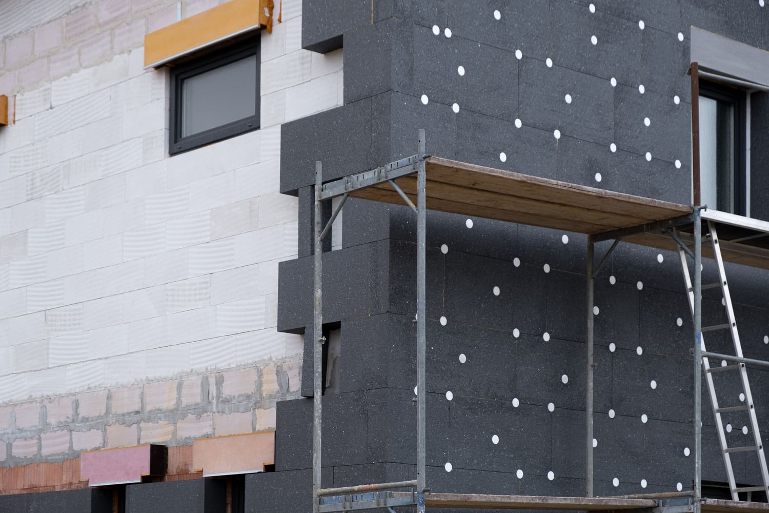 Construction site featuring a scaffold with black insulation panels being installed on a building's exterior, showcasing a mix of textured wall surfaces and white adhesive dots for securing the panels.