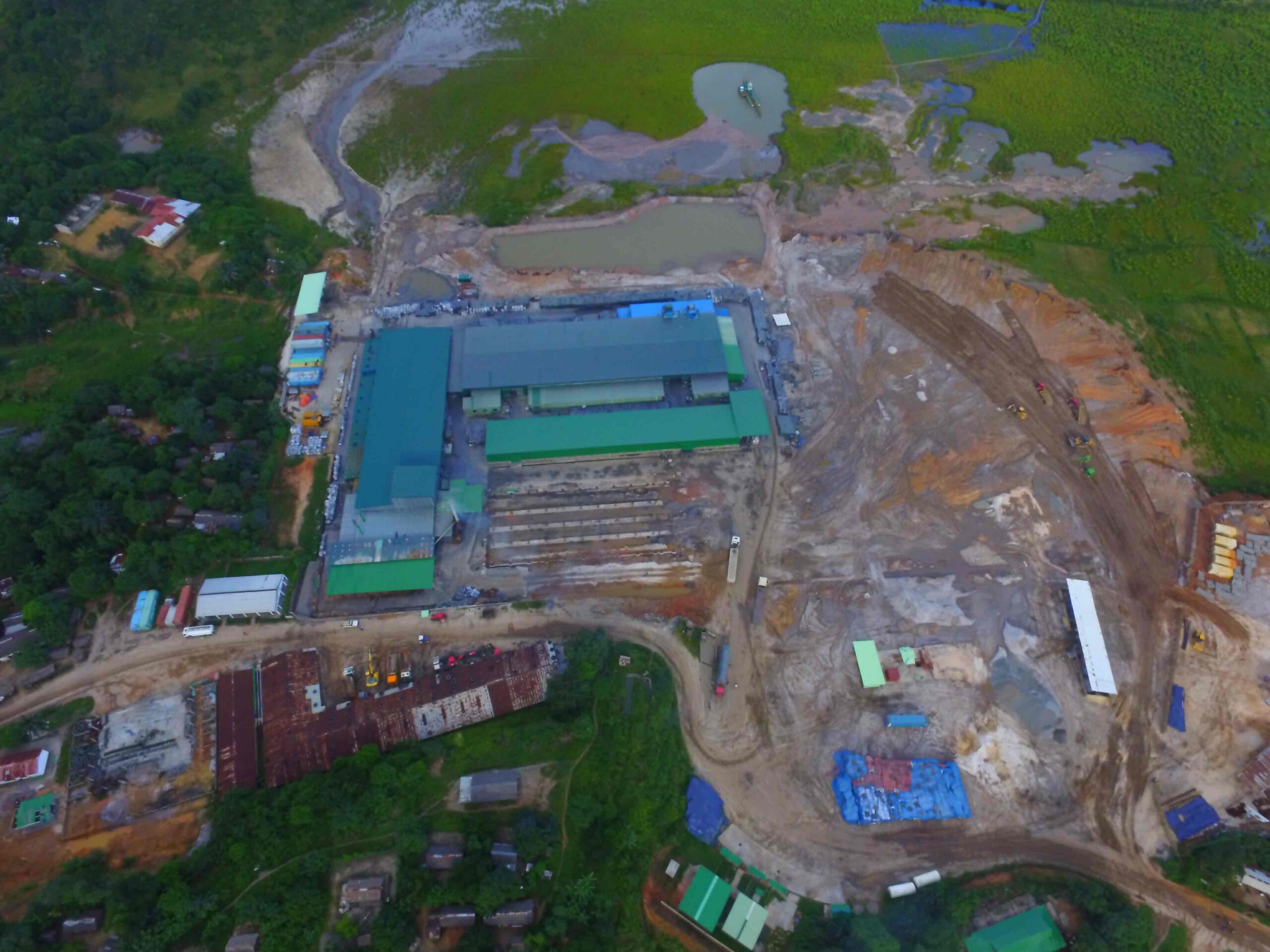 Aerial view of a construction site featuring large industrial buildings with green roofs, surrounded by cleared land, machinery, and various construction materials, set against a backdrop of lush greenery and water bodies.