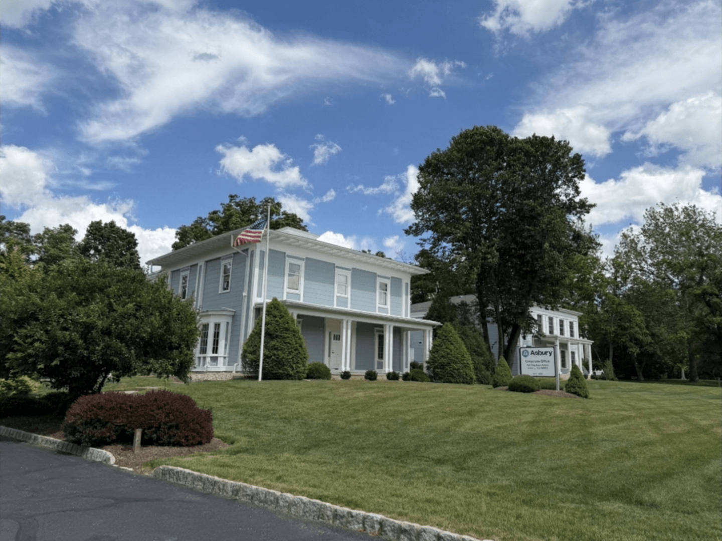 Two elegant houses with manicured lawns and trees, featuring an American flag, located at Asbury's corporate office. The sky is partly cloudy, creating a picturesque setting.